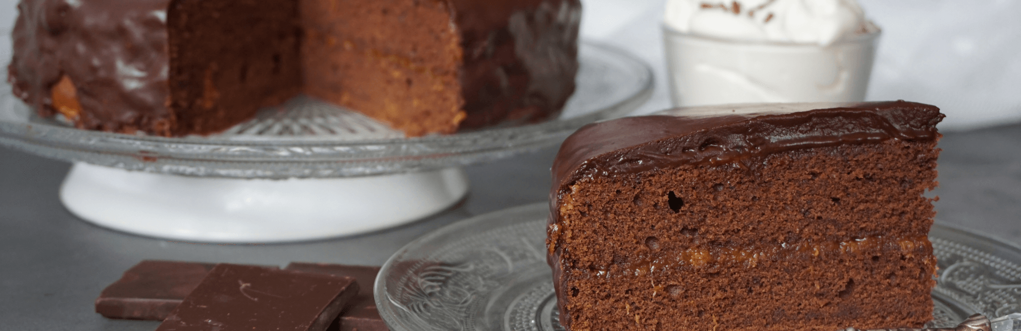 ein Stück Schokotorte auf einem Glasteller, im Hintergrund der Rest der Torte sowie ein Glas mit Schlagobers